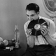1938: A man chews up a gramophone record. Beside him is a table on which are light bulbs waiting to be eaten.